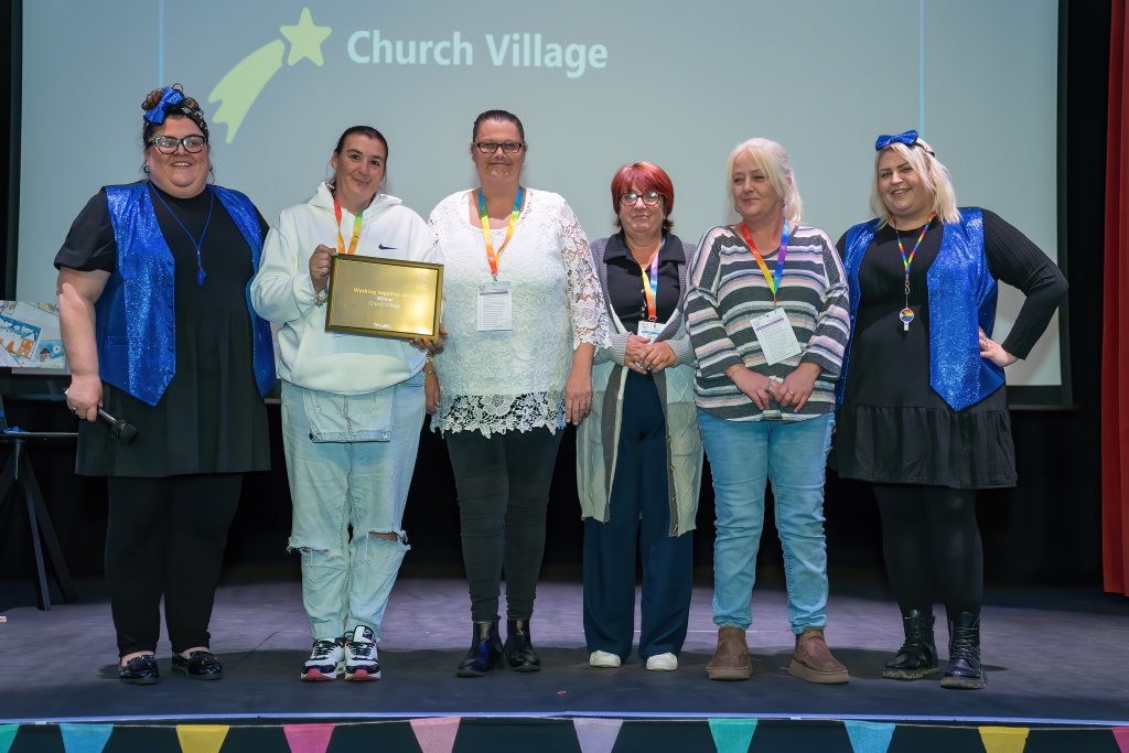 Trivallis Housing Landlord Wales Six people stand on a stage with a backdrop that reads "Church Village." One person holds a framed certificate. They are smiling and wearing casual attire with matching lanyards. A banner with colorful flags is visible at the bottom of the stage.