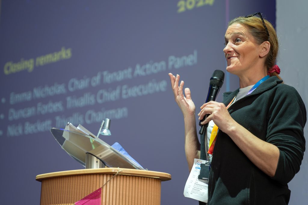 Trivallis Housing Landlord Wales A person stands at a podium giving a speech with a microphone. The background shows a projected slide listing closing remarks and names. The individual wears a lanyard and gestures with one hand while speaking.