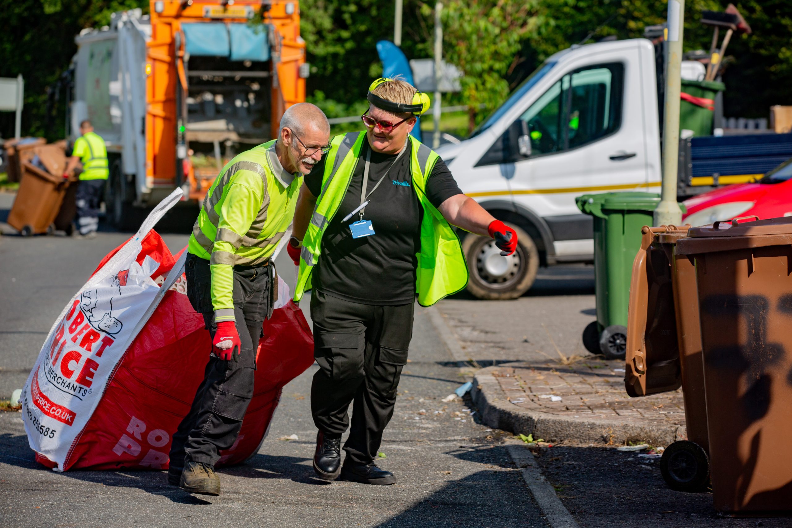 Trivallis Housing Landlord Wales Two sanitation workers, wearing safety vests and gloves, collaborate to handle large waste bags on a street. A garbage truck and another vehicle are visible in the background, along with several bins.