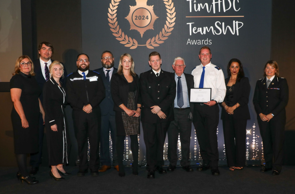 Trivallis Housing Landlord Wales A group of twelve people stand together on a stage, posing for a photo. One person holds a framed certificate. They are in front of a backdrop with the text "2024 Team HDC Team SWP Awards." Everyone is dressed in formal or uniform attire.