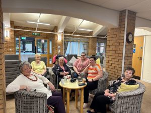 Trivallis Housing Landlord Wales A group of six older women sits together in a circle of armchairs in a cozy room. They are smiling and appear to be conversing. A small table in the center holds drinks and snacks. The room has brick walls, a clock, and large windows.