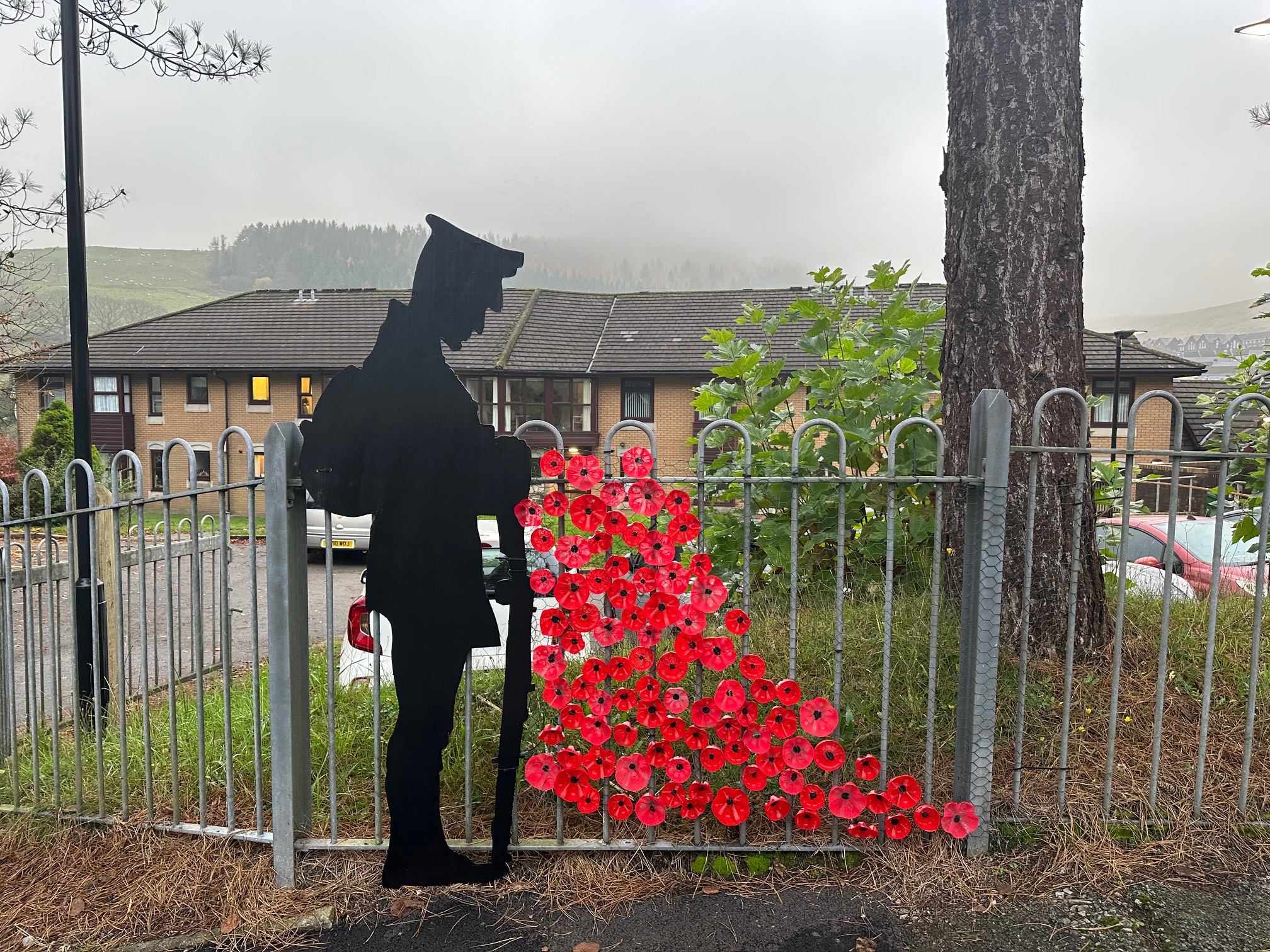 Trivallis Housing Landlord Wales Silhouette of a soldier stands against a metal fence with red poppies cascading down from the figure. A residential building and misty hills are in the background.