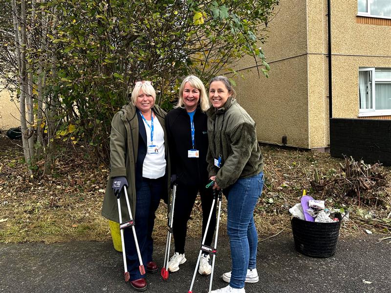 Trivallis Housing Landlord Wales Three women stand in front of a beige building, smiling at the camera. They are holding litter-picking tools and wearing ID badges. There is a basket filled with collected debris beside them, and foliage in the background.