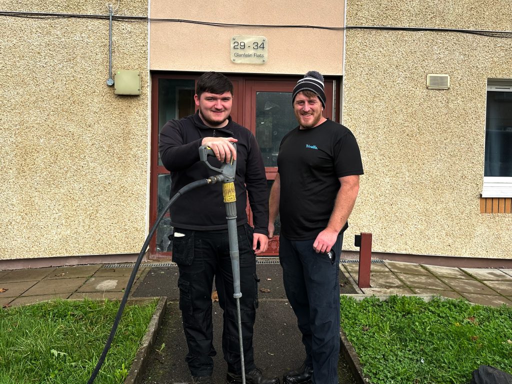 Trivallis Housing Landlord Wales Two men in work attire stand outside a building. One holds a tool with a hose attached. The entrance behind them has a sign reading "29-34 Guardian Flats." They are on a paved path with grass on either side.