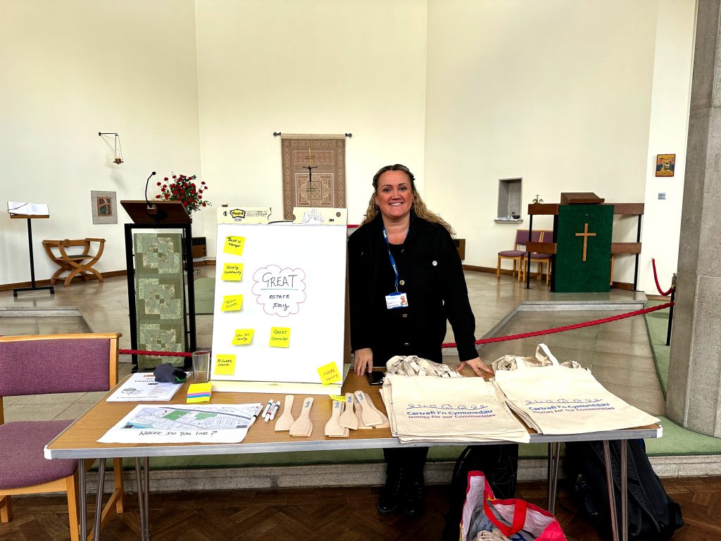 Trivallis Housing Landlord Wales A person stands behind a table in a church hall. The table has a whiteboard with sticky notes, informational materials, tote bags, and wooden utensils. A notice board, chairs, and a cross are visible in the background.