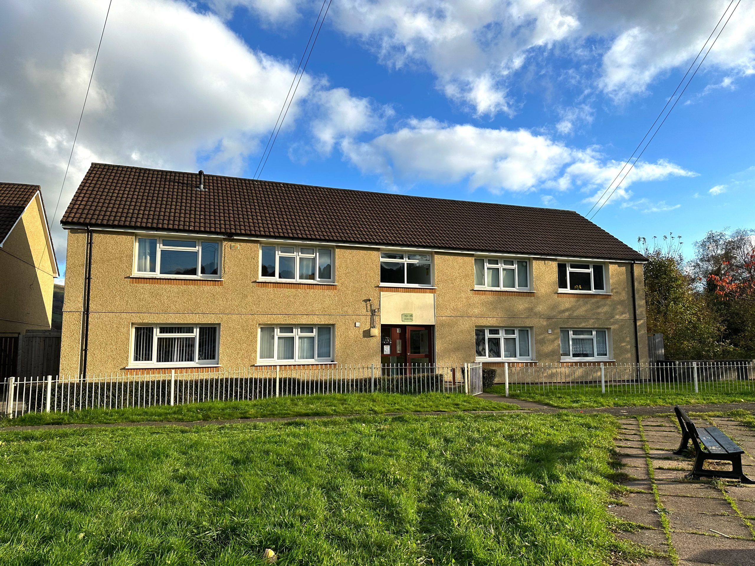 Trivallis Housing Landlord Wales A two-story residential building with a beige exterior and dark brown roof. The building has multiple white-framed windows. In front, there's a grassy area with a bench and a pathway. The sky is partly cloudy with patches of blue.