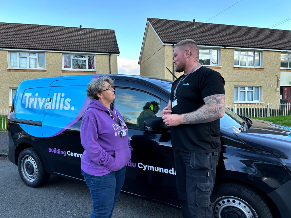 Trivallis Housing Landlord Wales A woman in a purple hoodie speaks with a man in a black shirt in front of a blue and black Trivallis van. They are standing on a residential street with houses and a cloudy sky in the background.