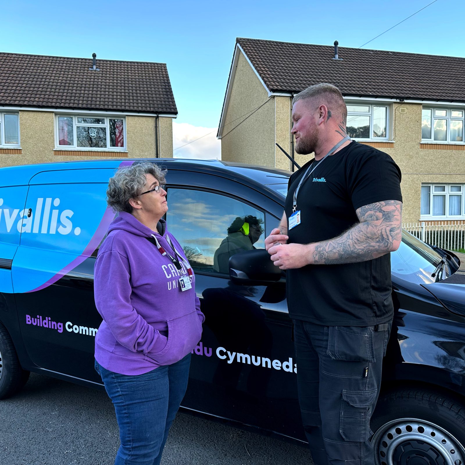 Trivallis Housing Landlord Wales A woman in a purple hoodie speaks with a man in a black shirt in front of a blue and black Trivallis van. They are standing on a residential street with houses and a cloudy sky in the background.