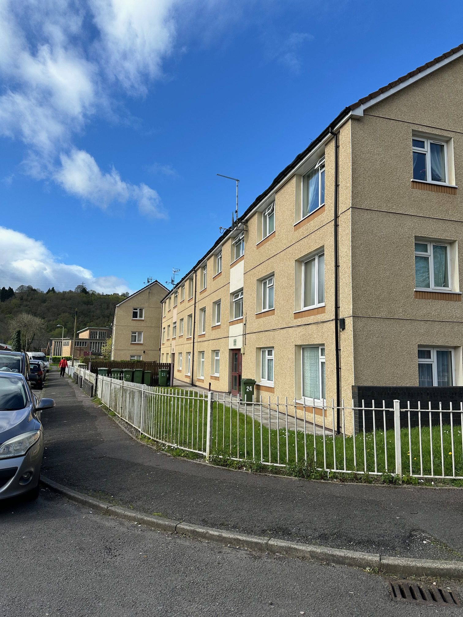 Trivallis Housing Landlord Wales View of a residential street with three-story apartment buildings on the right. Cars are parked along the left side of the road. A white metal fence borders the grass in front of the buildings. The sky is partly cloudy.