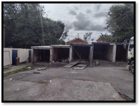 Trivallis Housing Landlord Wales Three empty garages with open doors are shown, with some debris scattered in front. A truck is partially visible on the right side. Trees and clouds fill the background. The scene appears overcast.