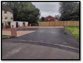 Trivallis Housing Landlord Wales Image of a newly paved driveway in a residential area. To the left, there are two brick structures with doors. A house and a fenced yard are visible in the background under a cloudy sky.