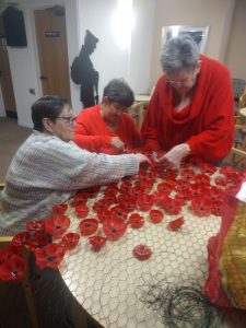 Trivallis Housing Landlord Wales Three people gather around a table, assembling red poppy flowers. The flowers are spread out on a surface covered with wire mesh. A shadow of a soldier is visible on the wall in the background.