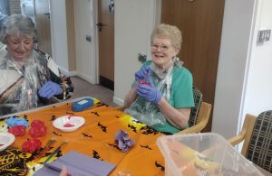 Trivallis Housing Landlord Wales Two older adults are sitting at a table covered with a Halloween-themed tablecloth, engaged in crafting activities. They are wearing plastic aprons and gloves. Decorations and craft supplies are on the table, with a ghost decoration hanging above.