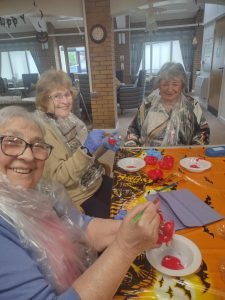 Trivallis Housing Landlord Wales Three elderly women are seated at a table decorated with an orange Halloween-themed tablecloth. They are painting items red and wearing plastic covers to protect their clothes. The room has festive decorations and a clock on a brick wall.