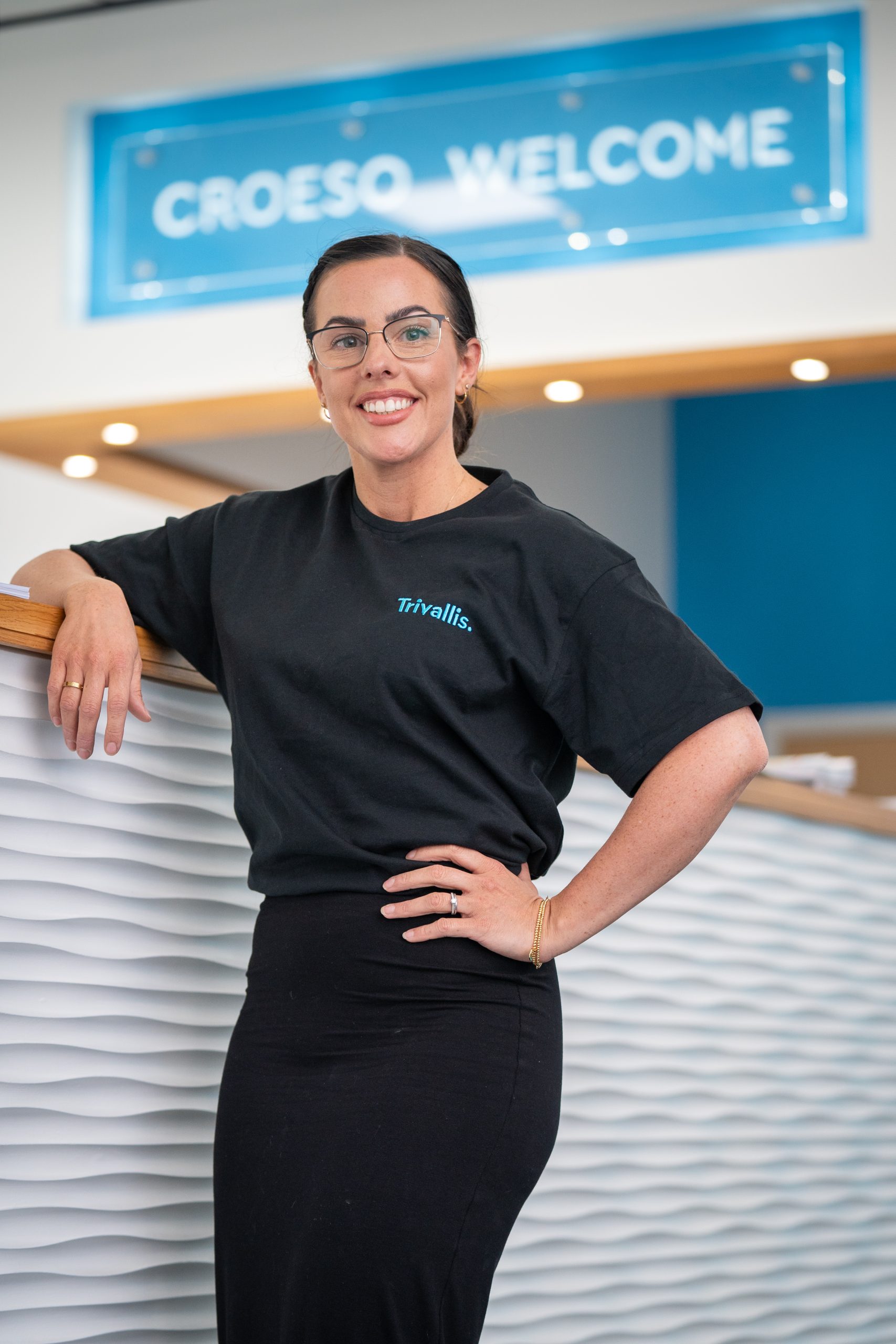 Trivallis Housing Landlord Wales A person wearing glasses and a black shirt stands smiling by a white and wood reception desk. Above, a blue sign reads "CROESO WELCOME.