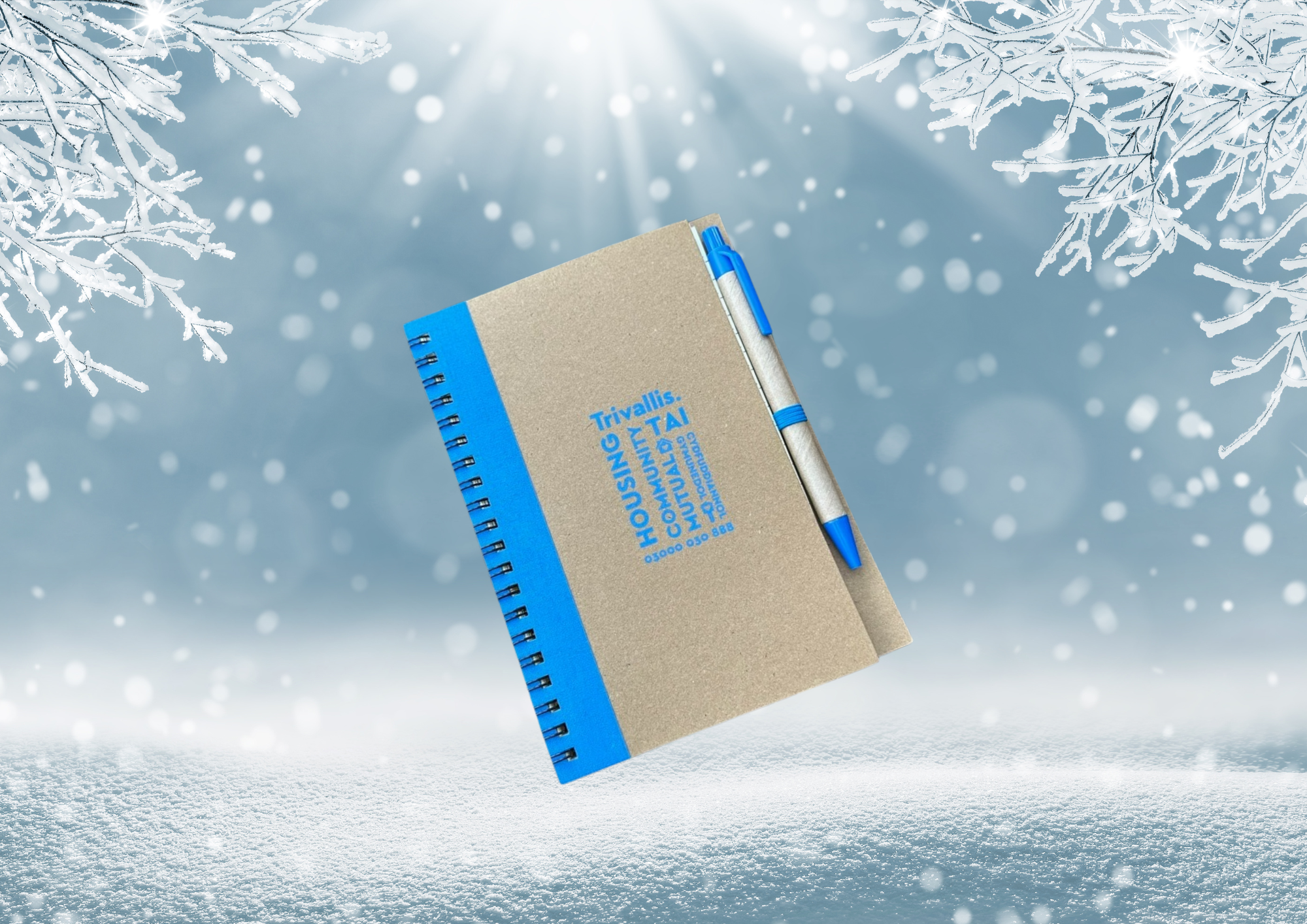 Trivallis Housing Landlord Wales A spiral-bound notebook with a brown cover and blue edges sits against a snowy background. A blue and silver pen is attached to the cover. Snowflakes and frosty branches are visible at the top corners of the image.