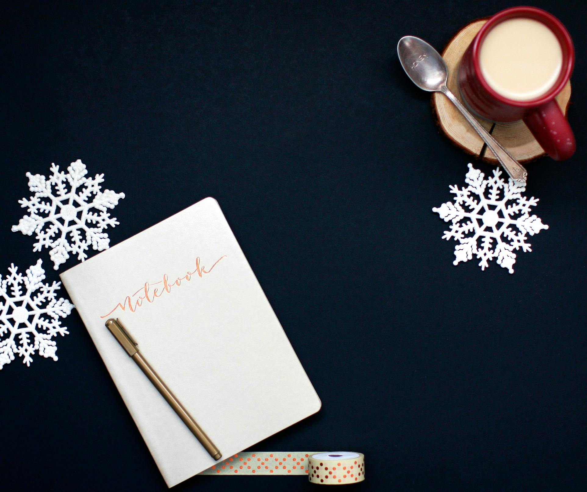 Trivallis Housing Landlord Wales A notebook with a pen rests on a dark surface next to two paper snowflakes. A red mug filled with a light-colored drink sits on a wooden coaster, accompanied by a spoon. Decorative tape is also visible.