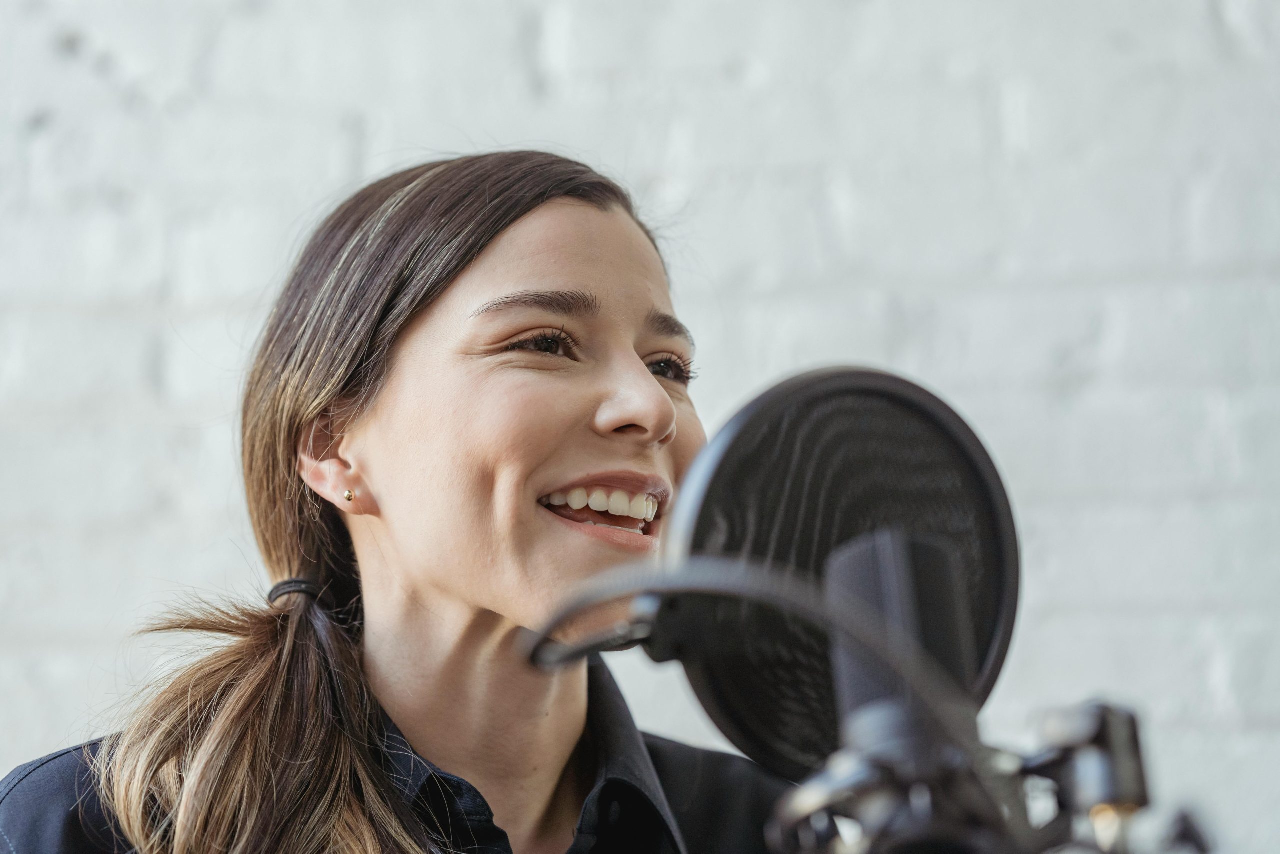 Trivallis Housing Landlord Wales A person with long hair and a ponytail smiles while speaking into a microphone with a pop filter. They are indoors, standing against a white brick wall.