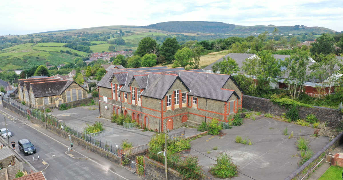 Trivallis Housing Landlord Wales An aerial view of an abandoned brick building with a pitched roof, surrounded by overgrown vegetation. The structure sits in a fenced area with a paved lot. Rolling hills and scattered houses are visible in the background under a cloudy sky.