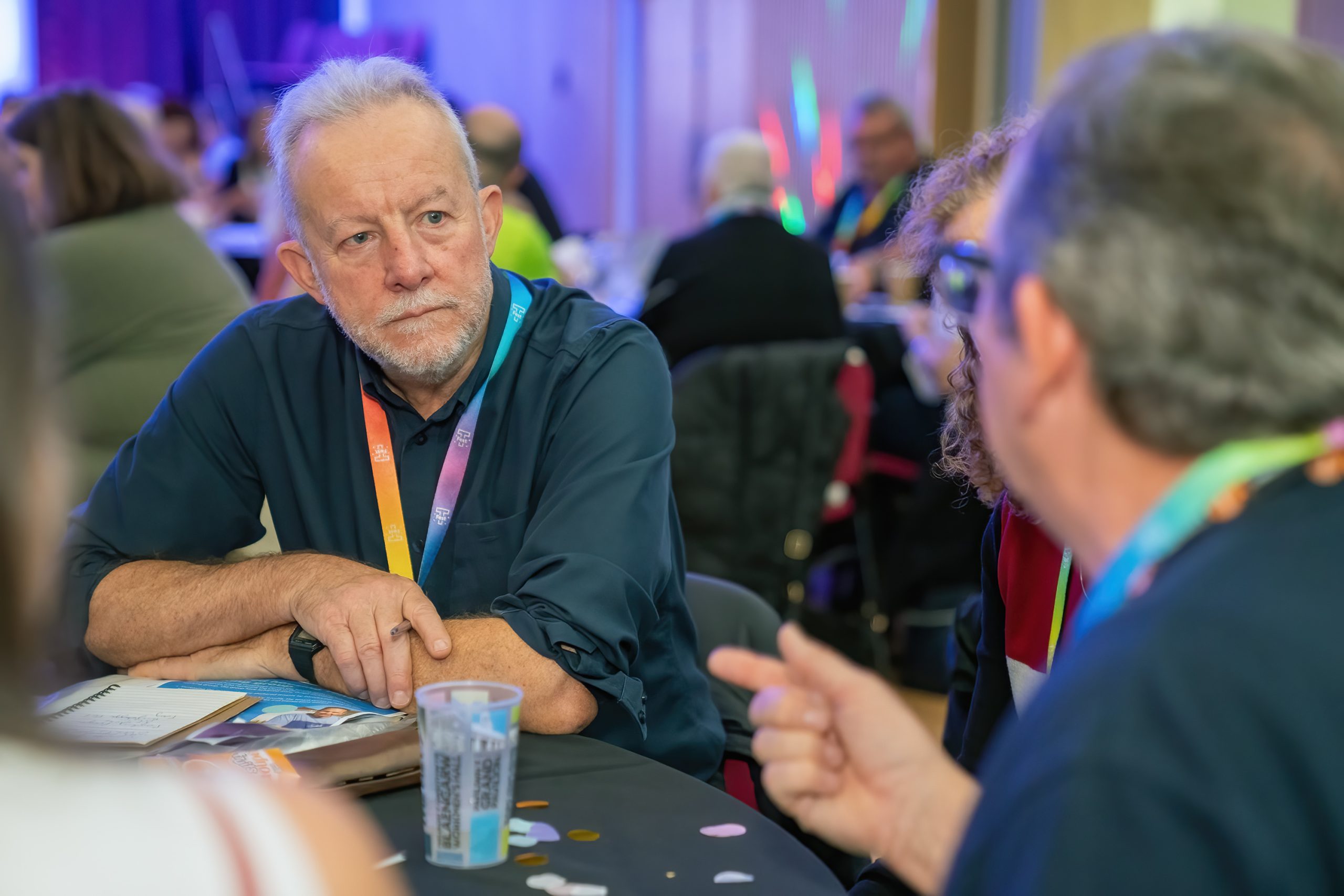 Trivallis Housing Landlord Wales A man with short gray hair and a beard, wearing a dark blue shirt, sits at a round table, engaging in conversation with others in a crowded room. He rests his elbow on the table and listens intently. The table has some papers and a plastic cup.