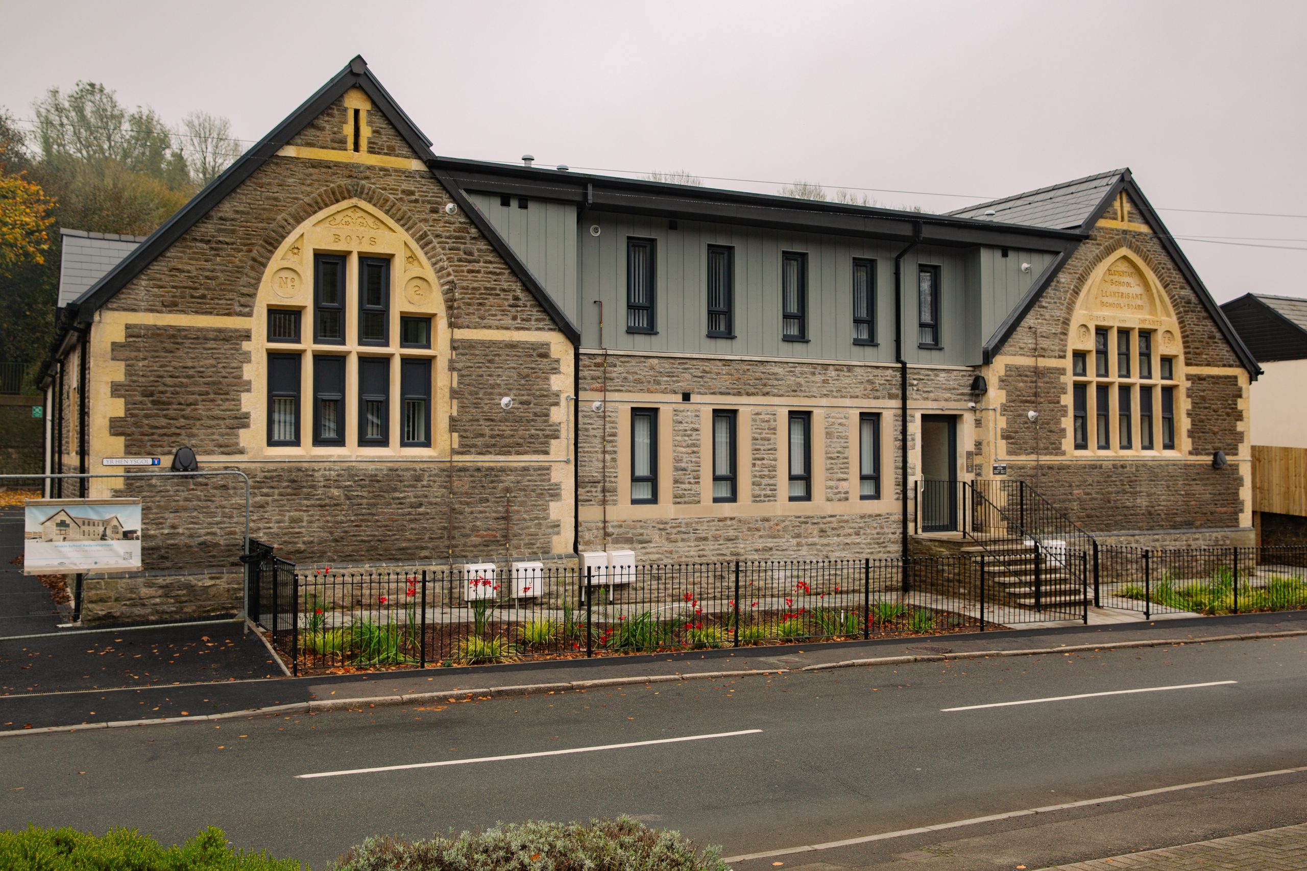 Trivallis Housing Landlord Wales A modern building with stone and beige exterior, featuring large arched windows. It has a gabled roof and is surrounded by a black metal fence. A banner is displayed outside. The road in front is empty, and there are trees in the background.