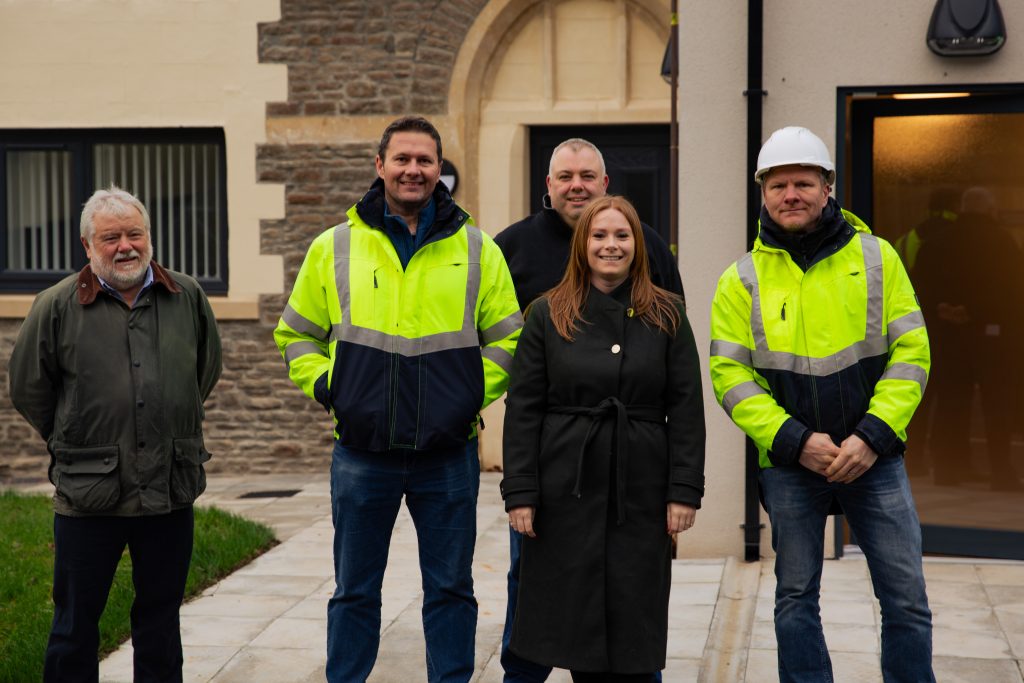 Trivallis Housing Landlord Wales Five people are standing outdoors on a paved area. Two individuals wear yellow safety jackets, and one person wears a white hard hat. The group appears in front of a building made of brick and stone. They are all smiling at the camera.