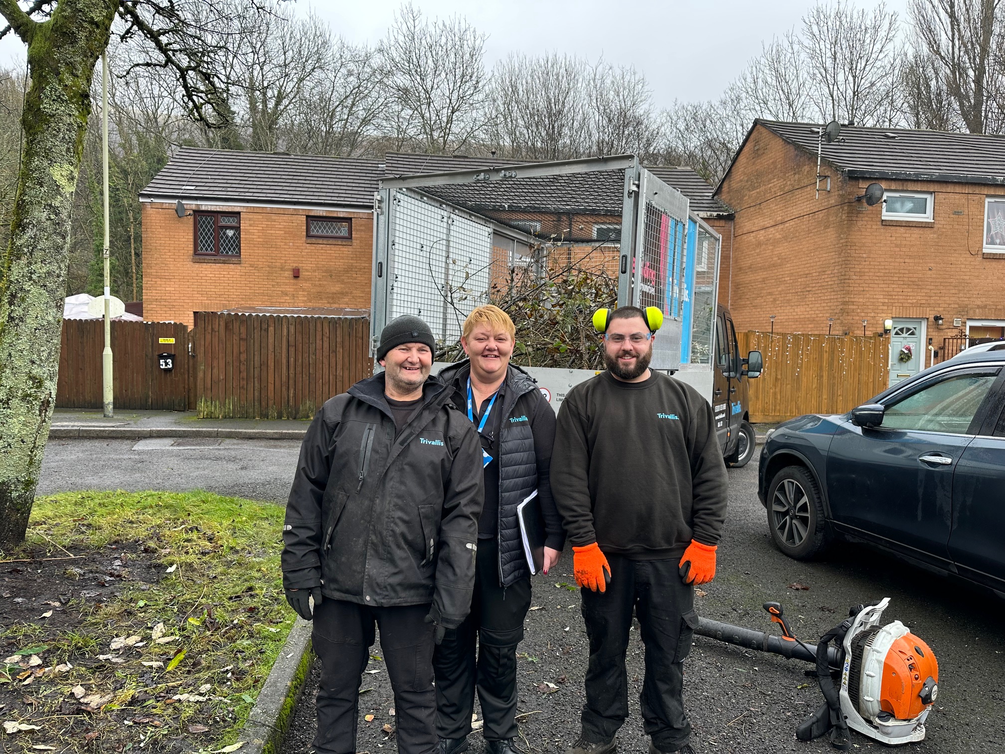 Trivallis Housing Landlord Wales Three people are standing outdoors in front of a piece of machinery. Two men are wearing black work clothes and gloves; one also has ear protection. A woman in a black jacket stands between them. Houses and trees are visible in the background.