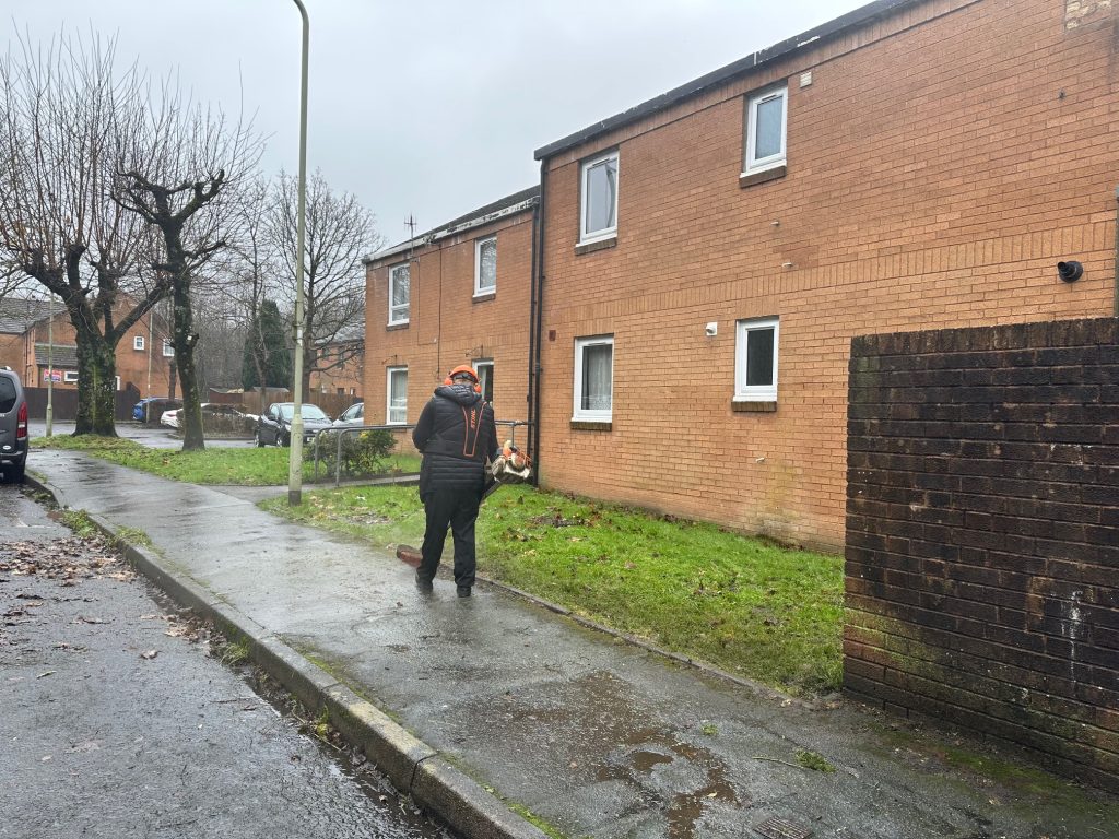 Trivallis Housing Landlord Wales Person using a leaf blower on a wet pavement next to a grass patch and brick building. It's a cloudy day, and trees without leaves line the walkway. A parked vehicle is partially visible in the distance.