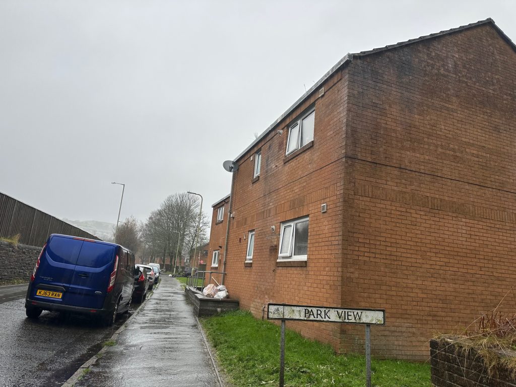 Trivallis Housing Landlord Wales A brick residential building is situated along a wet street. The road is lined with parked vehicles, including a blue van. A street sign reads "Park View." The sky is overcast, and the trees are bare.