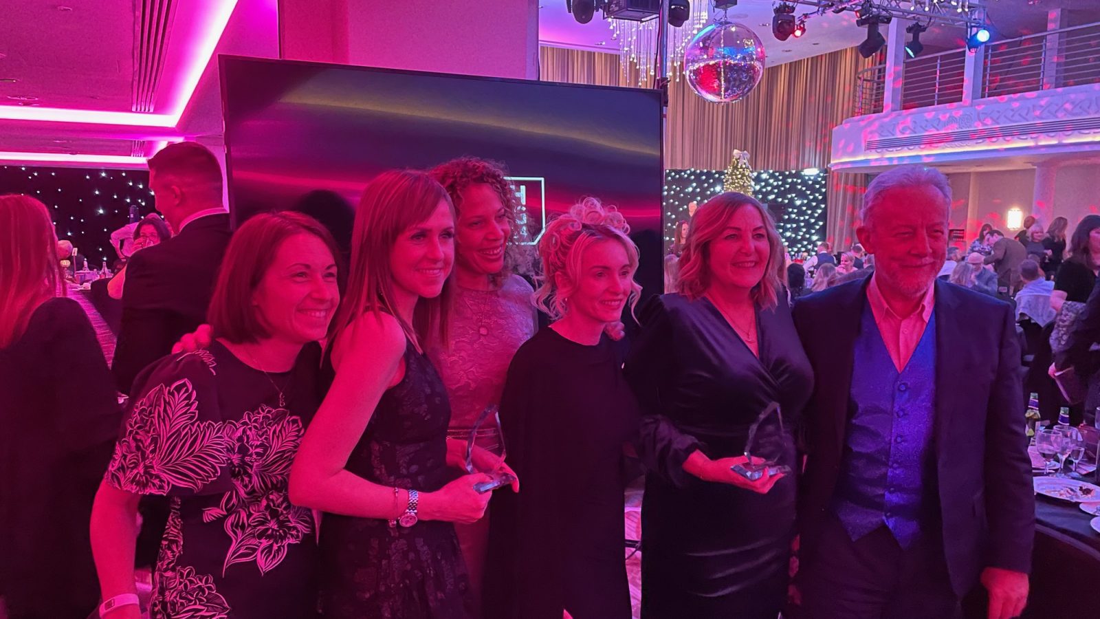 Trivallis Housing Landlord Wales A group of people dressed in formal attire are posing together in a banquet hall. The room is dimly lit with pink and blue lighting, and there are decorations like a disco ball and chandeliers. The atmosphere appears to be celebratory.