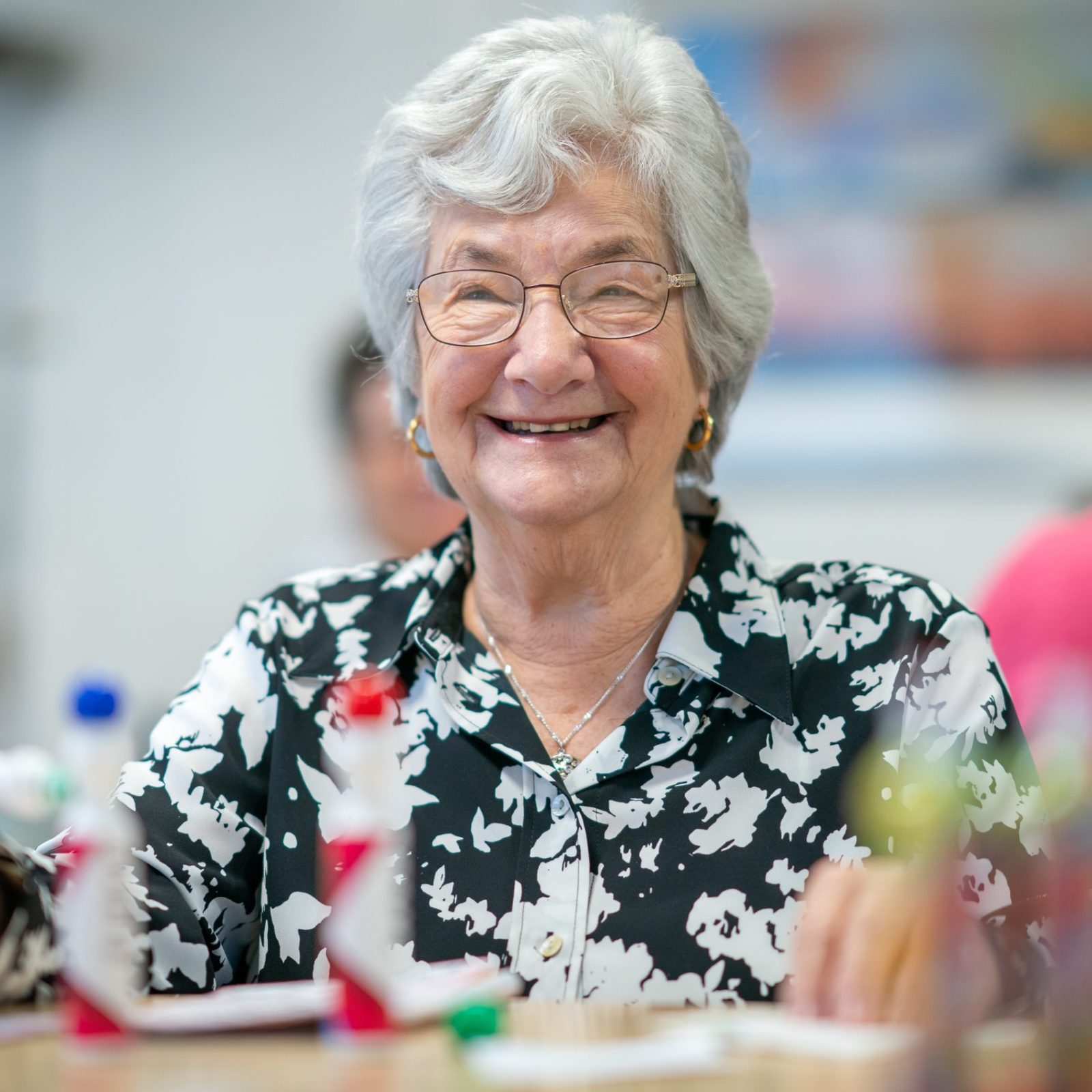 Trivallis Housing Landlord Wales An elderly woman with gray hair and glasses is smiling while holding a bingo marker in a room with blurred people in the background. She is wearing a black and white patterned blouse.