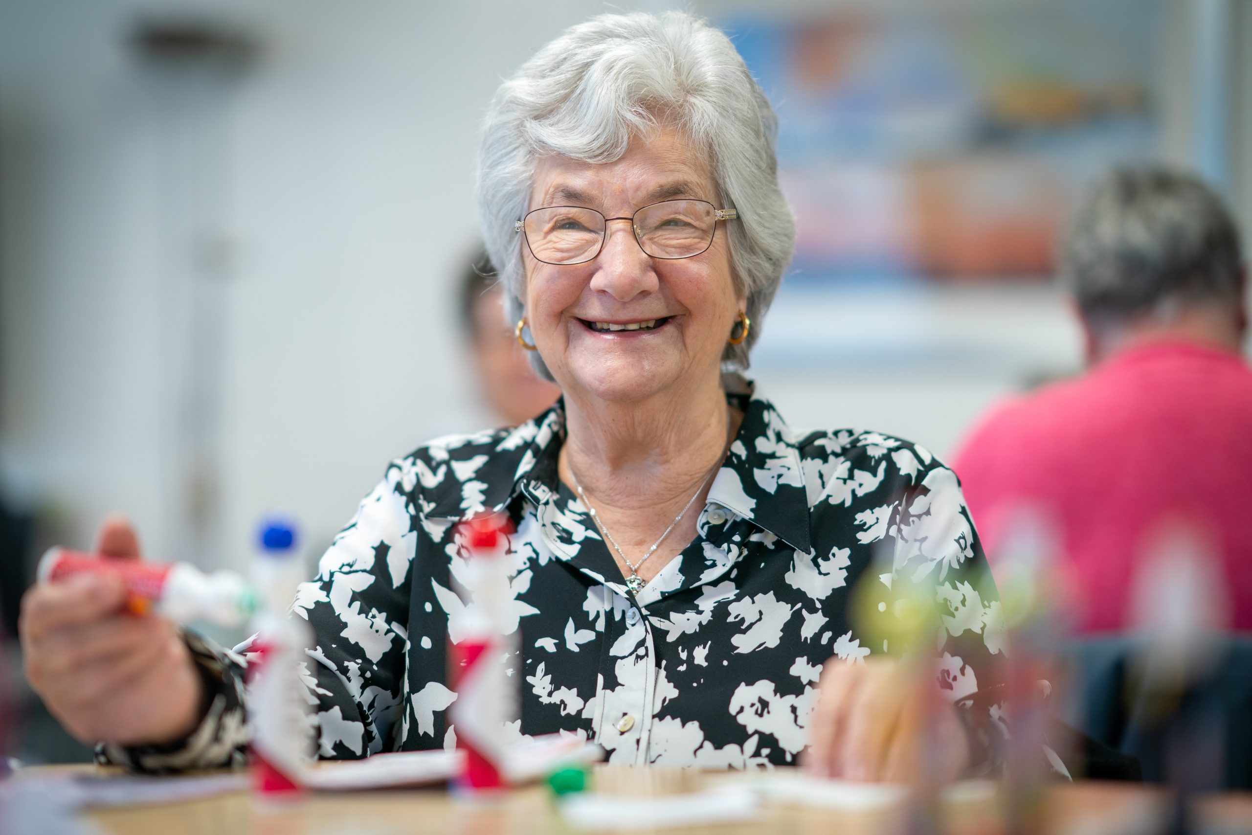 Trivallis Housing Landlord Wales An elderly woman with gray hair and glasses is smiling while holding a bingo marker in a room with blurred people in the background. She is wearing a black and white patterned blouse.