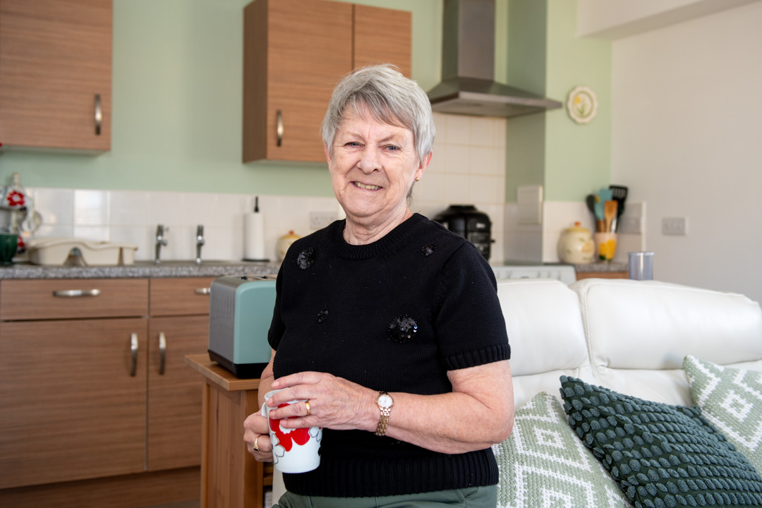 Trivallis Housing Landlord Wales An elderly woman with short gray hair is standing in a kitchen, holding a mug. She is wearing a black sweater and green pants. The kitchen has wooden cabinets, a toaster, and various kitchen utensils on the counter. A patterned cushion is on a white sofa.