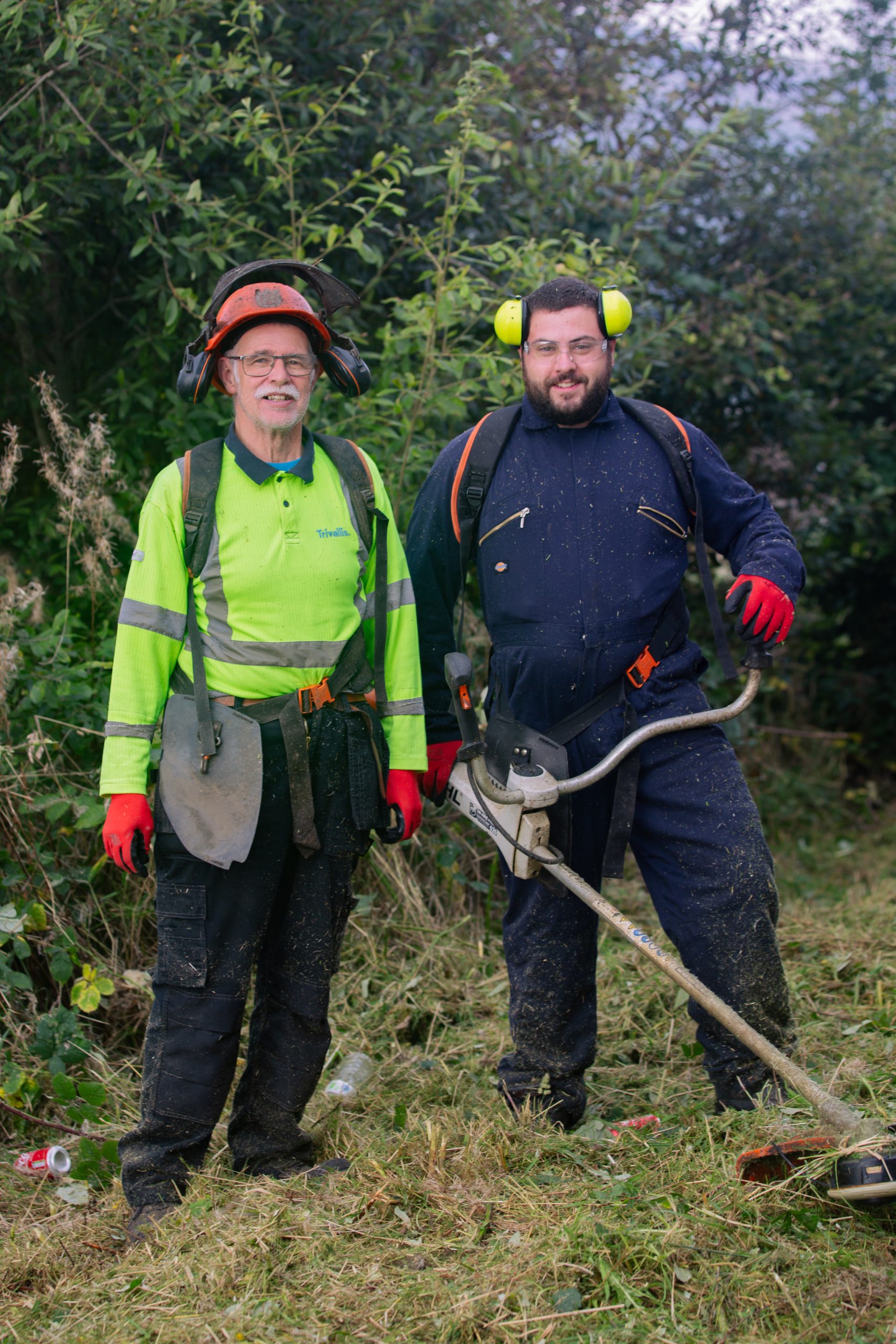 Trivallis Housing Landlord Wales Two people in protective gear stand outdoors on grass. One wears a lime green safety helmet and harness, the other has ear protection and a trimmer. Dense bushes and trees are in the background.