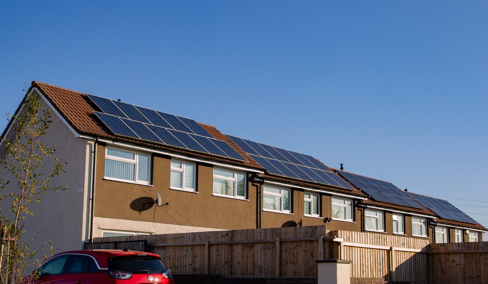 Trivallis Housing Landlord Wales A row of houses with solar panels on the roofs under a clear blue sky. A red car is parked on the driveway in front of a wooden fence.