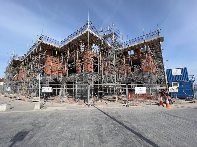 Trivallis Housing Landlord Wales A large building under construction with scaffolding covering its exterior. The brick structure is partially completed. Construction signs and cones are situated around the site, and a blue portable cabin is visible on the right. Clear sky overhead.