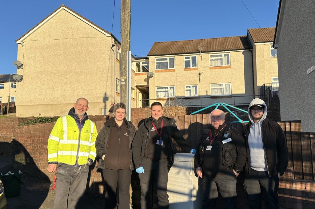 Trivallis Housing Landlord Wales A group of five people, including a person in a high-visibility jacket, standing on a residential street in front of houses under a clear blue sky. There is a trash bin nearby.