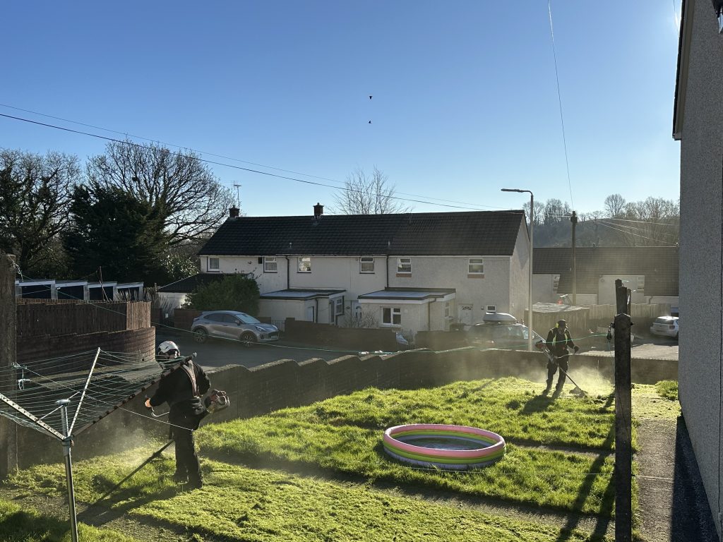 Trivallis Housing Landlord Wales Two people are using grass trimmers on a sunny lawn. A small, colorful inflatable pool sits on the grass. Houses and parked cars are visible in the background, with a blue sky overhead.