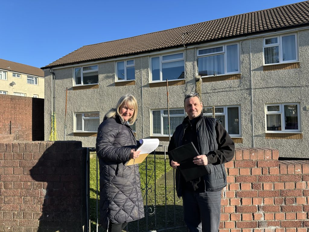 Trivallis Housing Landlord Wales Two individuals stand outside in front of a two-story residential building. Both are holding papers or notebooks. The woman wears a long dark coat, and the man has a dark jacket. They are next to a brick wall and iron gate. The sky is clear and blue.