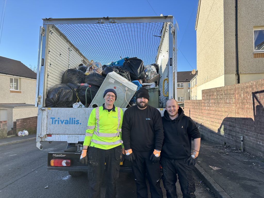 Trivallis Housing Landlord Wales Three people stand in front of a garbage truck filled with black bags and other refuse. They wear work clothes, including gloves, and are on a residential street. The truck has "Trivallis" written on it. The background shows houses and a brick wall.