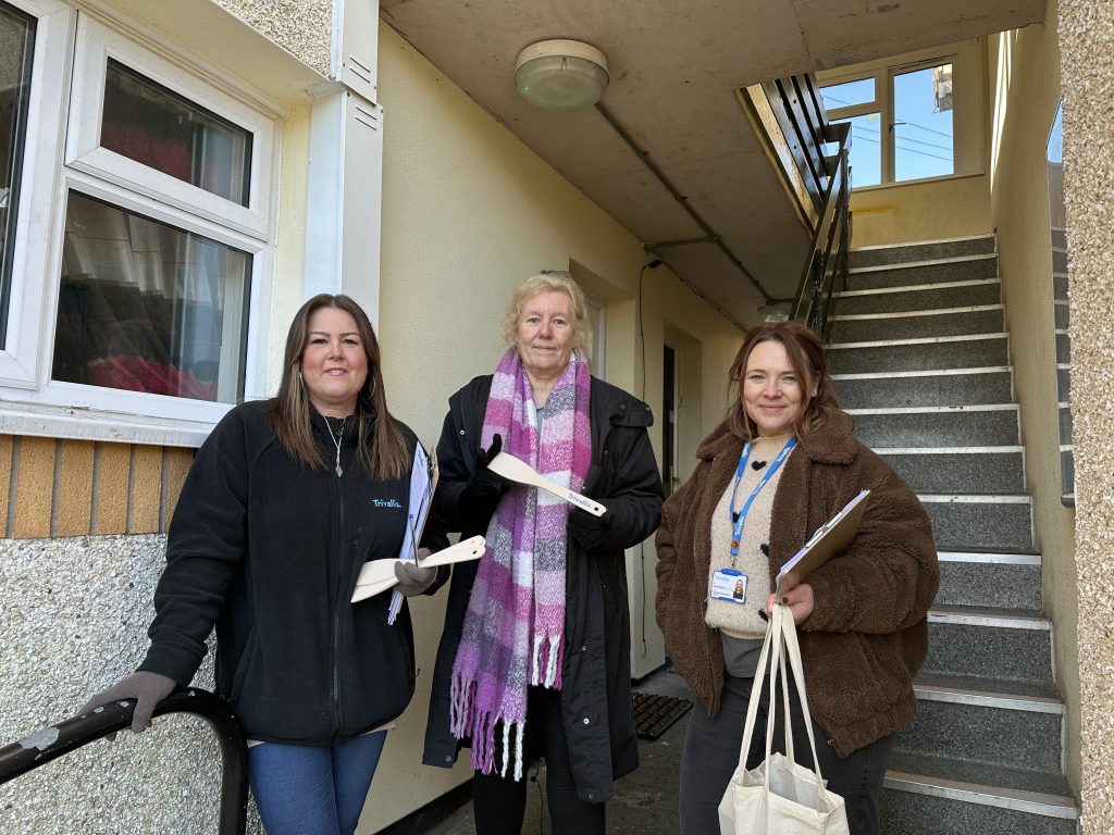 Trivallis Housing Landlord Wales Three women stand in a corridor with a staircase behind them. They each hold papers and one carries a bag. Two are wearing coats, and the middle one has a scarf. The setting appears to be a residential building.