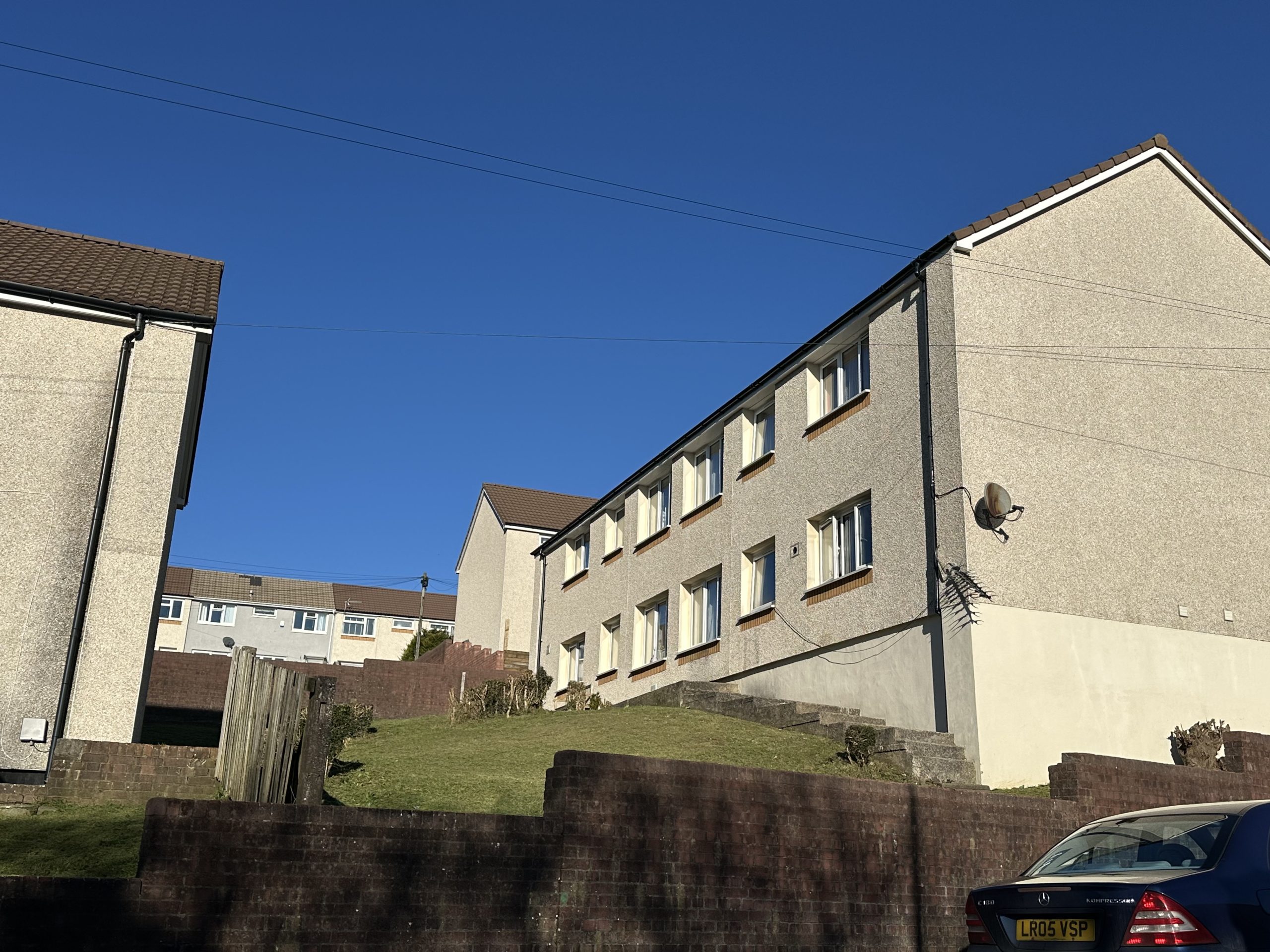 Trivallis Housing Landlord Wales A two-story residential building with plain white walls and multiple windows. It is situated on a sloping grassy area with a brick retaining wall. A parked car is partially visible on the right. The sky is clear and blue.