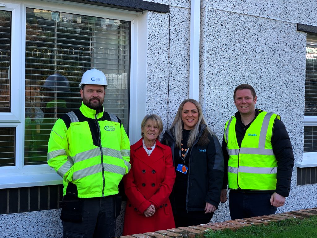 Trivallis Housing Landlord Wales Four people stand in front of a building with a window. Two men wear reflective vests, one with a hard hat. A woman in a red coat and another in a black jacket are between them. They are outdoors on a sunny day.