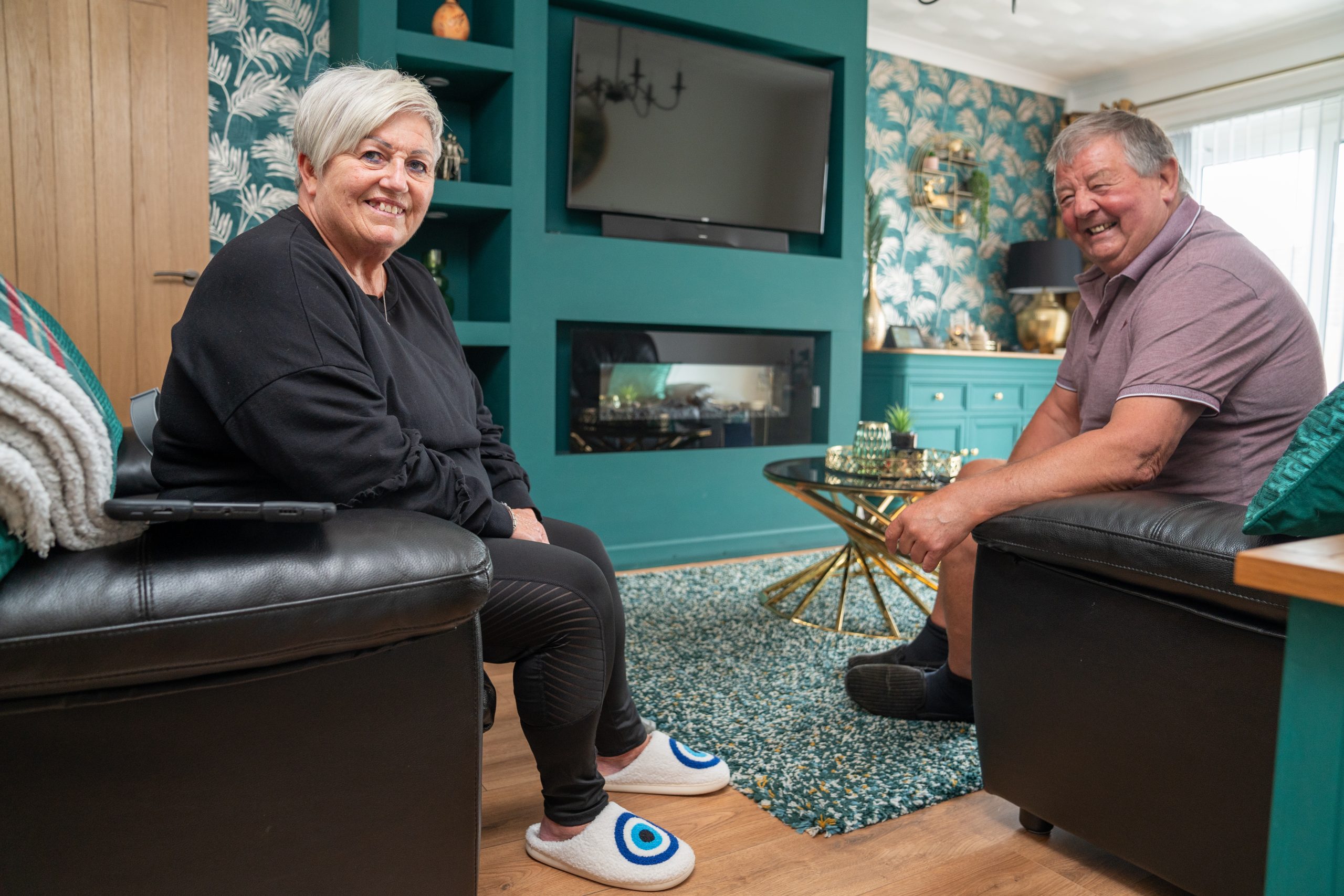 Trivallis Housing Landlord Wales A woman and a man are sitting in a living room, facing each other and smiling. The room has a green accent wall with floral wallpaper, a flat-screen TV, and a fireplace. A round coffee table is in the center with decorative items on it.