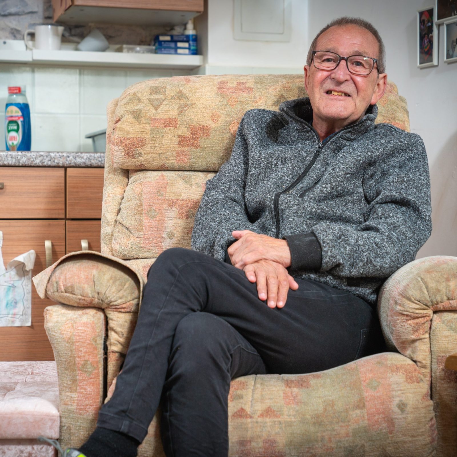 Trivallis Housing Landlord Wales An older man with glasses is sitting comfortably on a patterned armchair in a small kitchen-living area. He is wearing a gray zip-up jacket and black pants, with his legs crossed and hands resting in his lap. There's a footstool nearby.