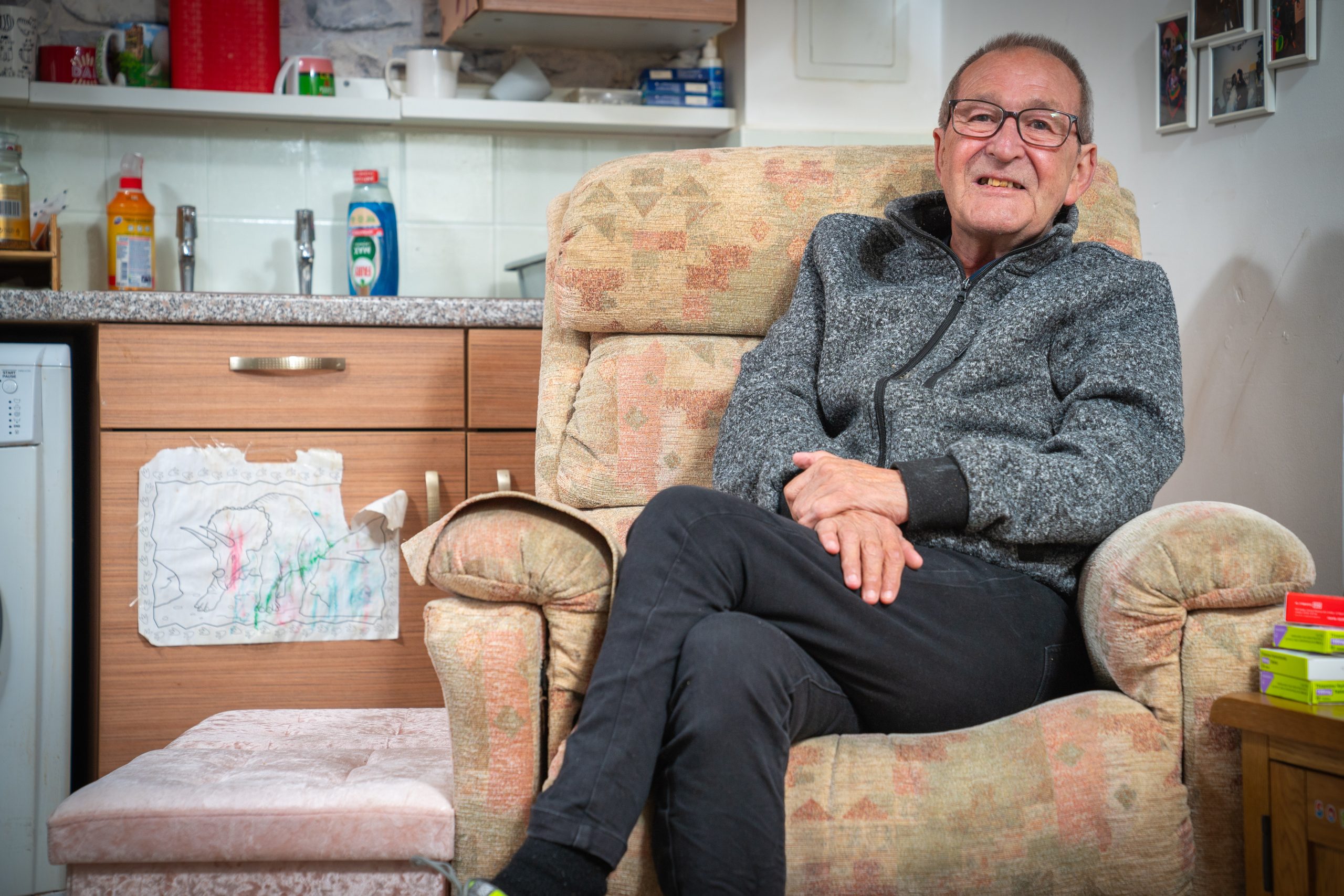 Trivallis Housing Landlord Wales An older man with glasses is sitting comfortably on a patterned armchair in a small kitchen-living area. He is wearing a gray zip-up jacket and black pants, with his legs crossed and hands resting in his lap. There's a footstool nearby.