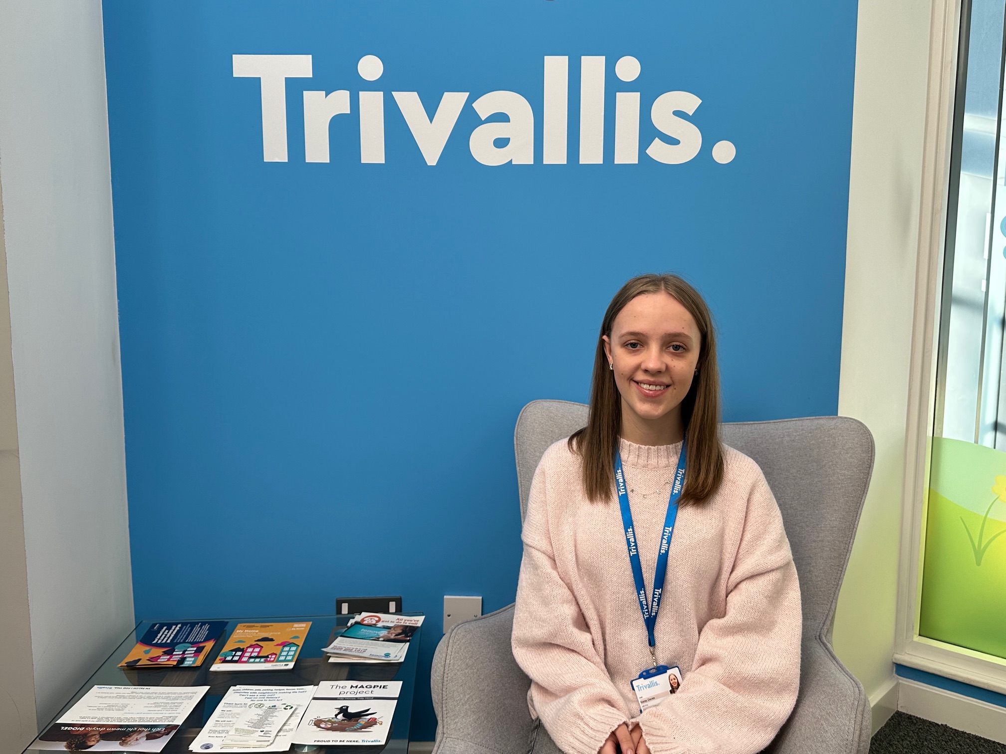 Trivallis Housing Landlord Wales A person with long hair sits in a gray chair wearing a pink sweater and a lanyard with an ID badge. Behind them is a blue wall with the word "Trivallis." A table nearby holds brochures and magazines.