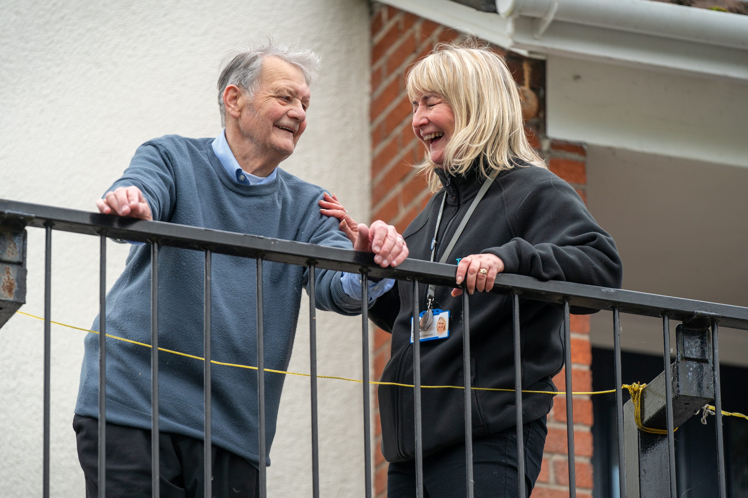 Trivallis Housing Landlord Wales An elderly man and a woman stand on a balcony smiling at each other. The man is wearing a blue sweater, and the woman is wearing a black jacket with an ID badge. They appear to be engaged in a friendly conversation.