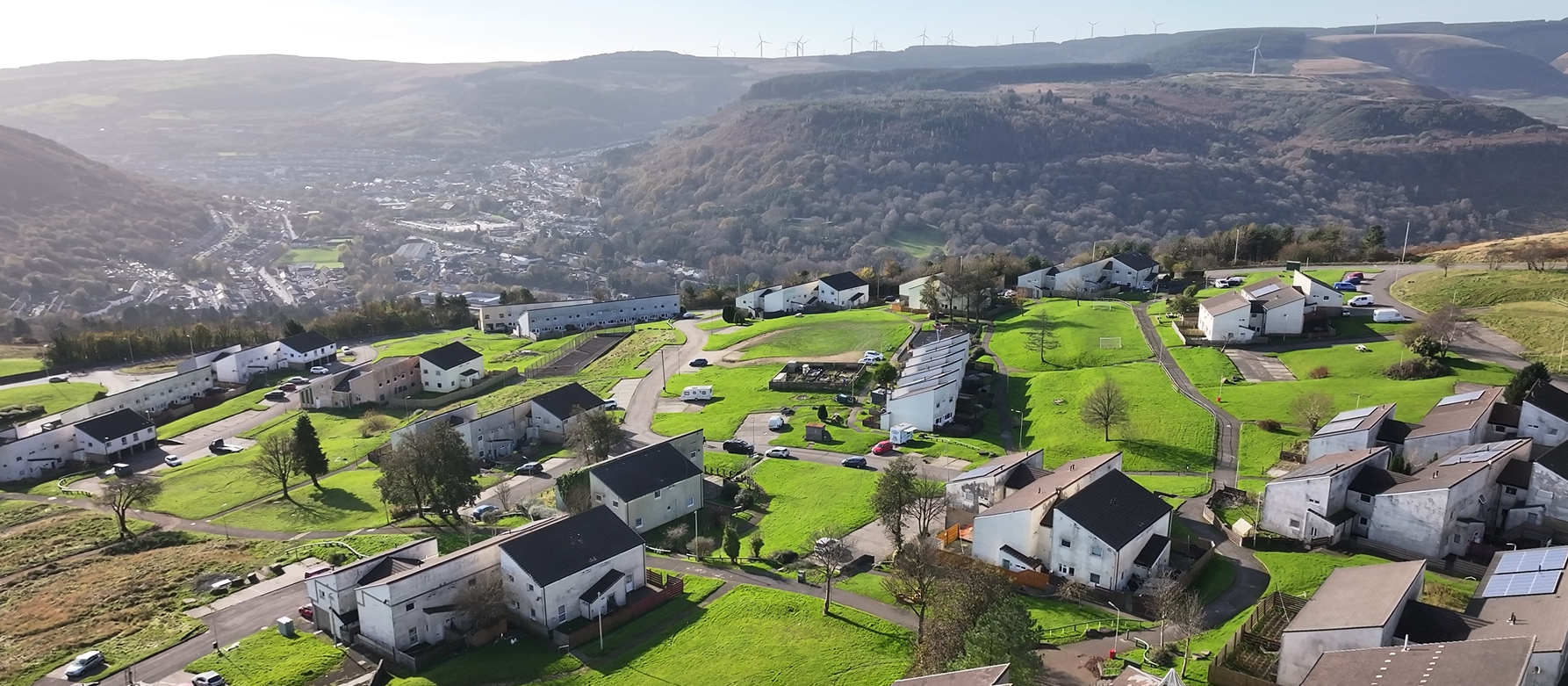 Trivallis Housing Landlord Wales Aerial view of a small hillside village with clusters of white houses, surrounded by green lawns and scattered trees. In the background, a town is nestled in a valley with rolling hills and wind turbines visible on the horizon.