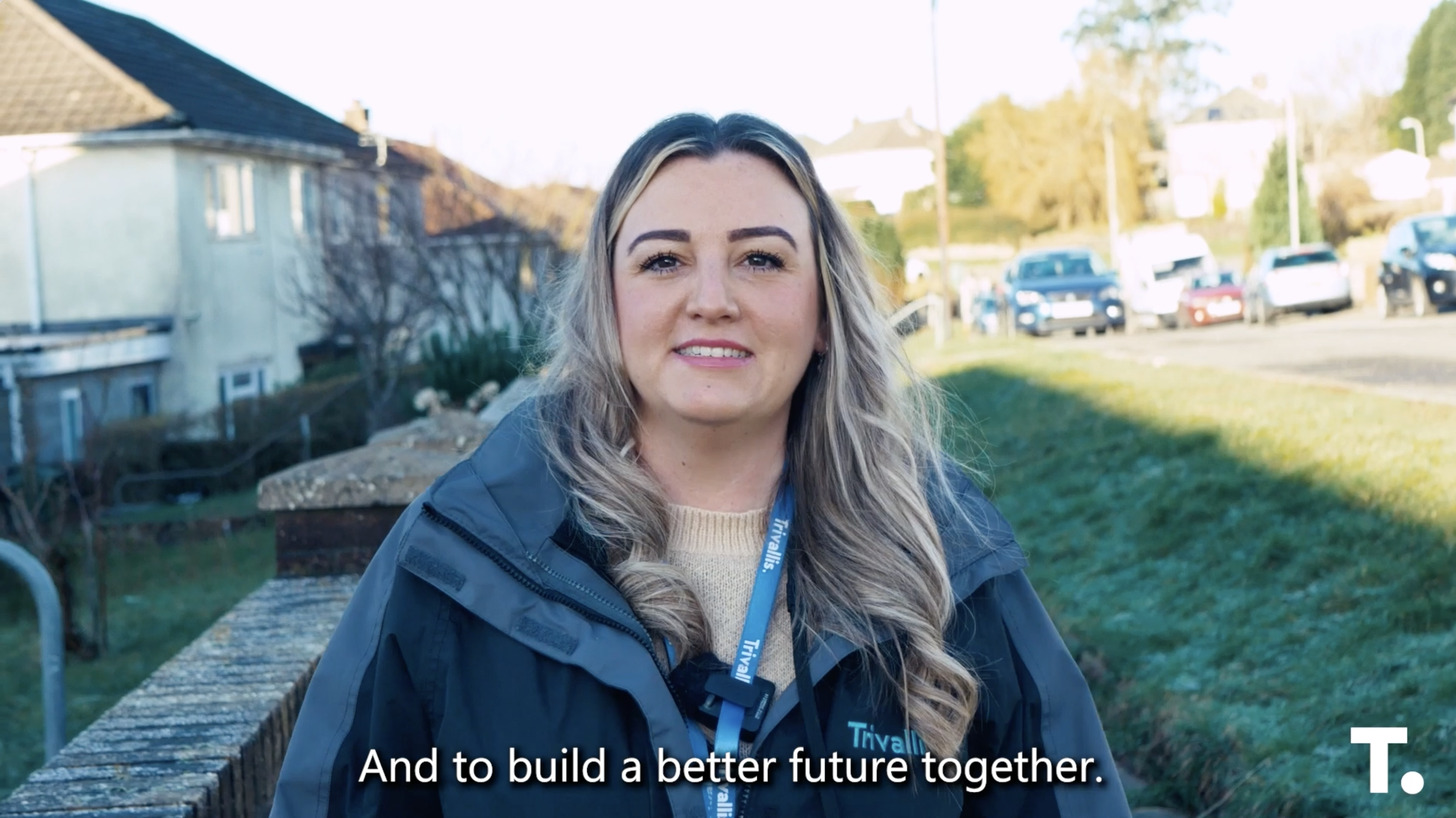 Trivallis Housing Landlord Wales A woman wearing a gray jacket stands outdoors, smiling at the camera. She has long hair and a lanyard around her neck. Houses, trees, and cars are visible in the background. The subtitle reads, "And to build a better future together.